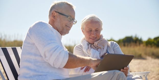 Due anziani su una sdraio in spiaggia, intenti a usare un tablet, godendosi il loro tempo libero in pensione.
