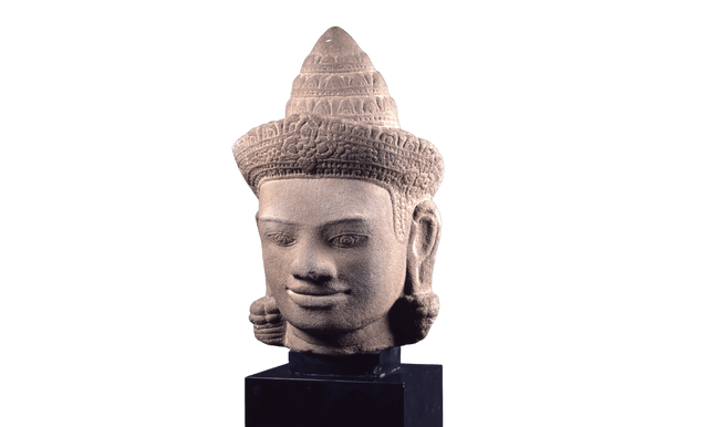 Statua raffigurante una testa di divinità, caratterizzata da un sorriso lievemente abbozzato e dal copricapo a forma di cono con decorazioni incise.