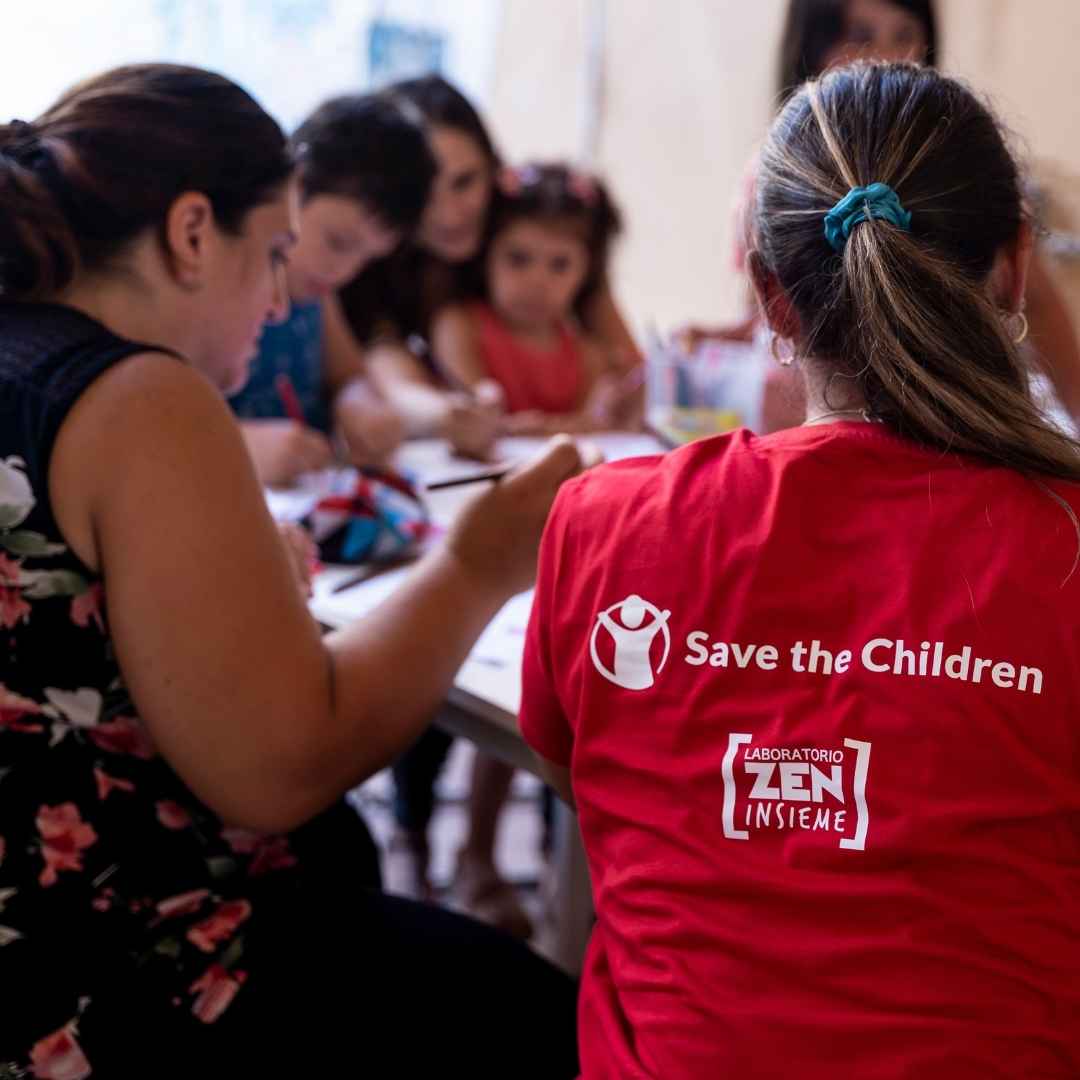 Operatrice di Save the Children in maglia rossa, seduta a un tavolo, mentre segue un'attività di doposcuola a bambini.